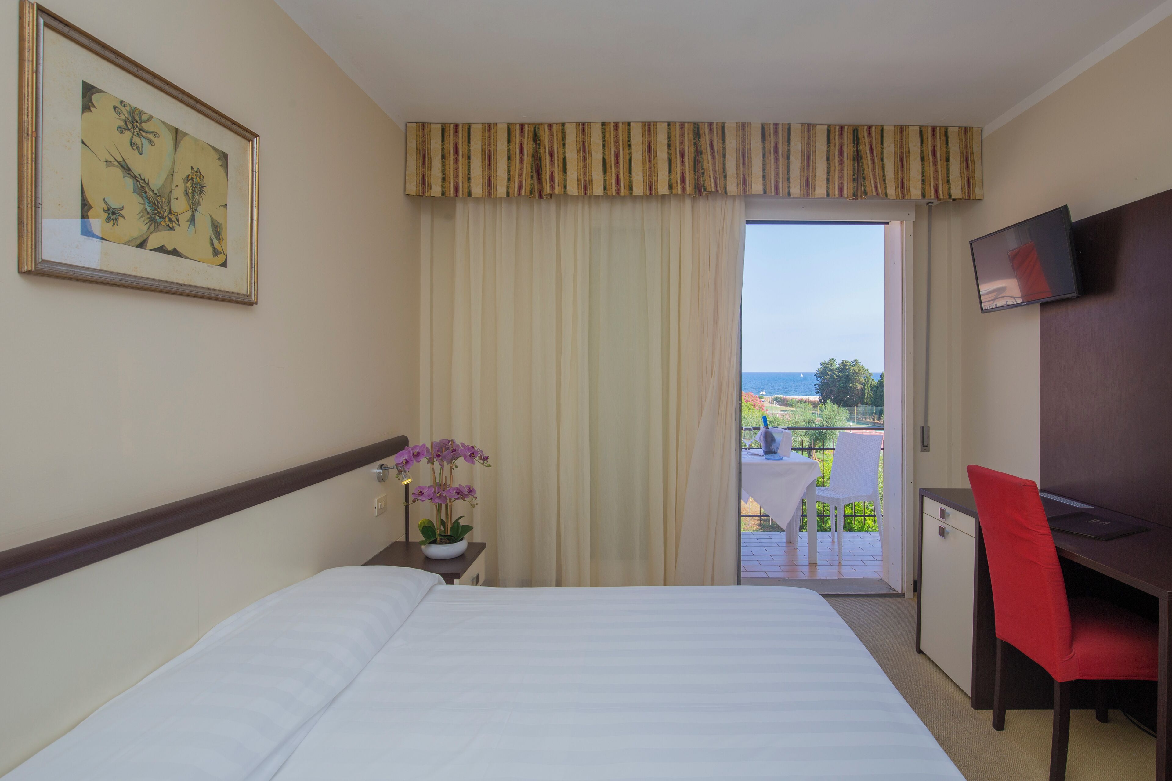 Habitación de hotel con cama, escritorio, silla, televisor, ventana con cortinas y un cuadro en la pared.