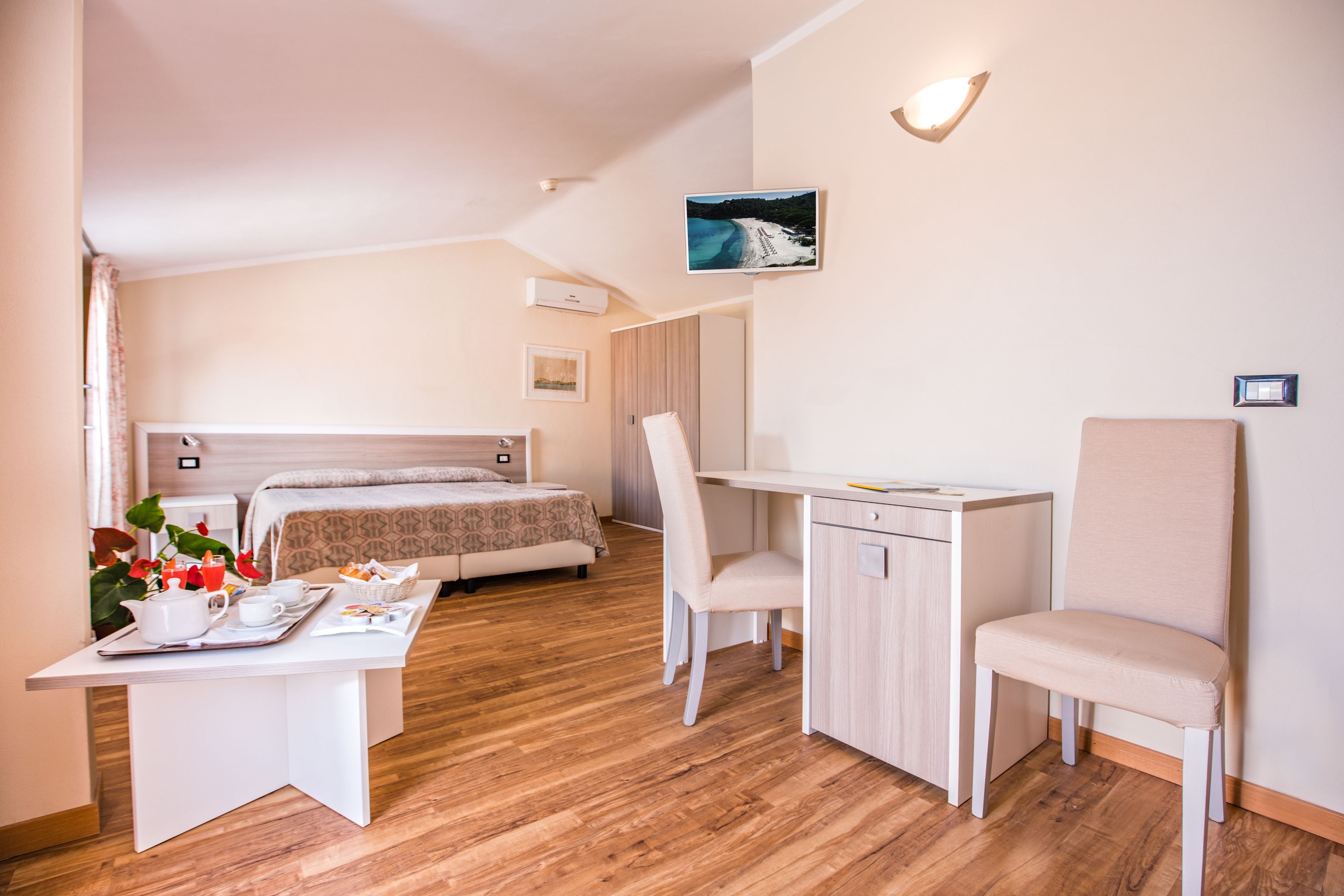 A modern hotel room with a bed, desk, and chair, a small table with breakfast items, and a picture on the wall.