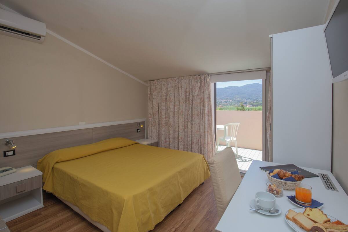 A hotel room with a bed, a bedside table, a small table with breakfast items, and a view of the outdoors.