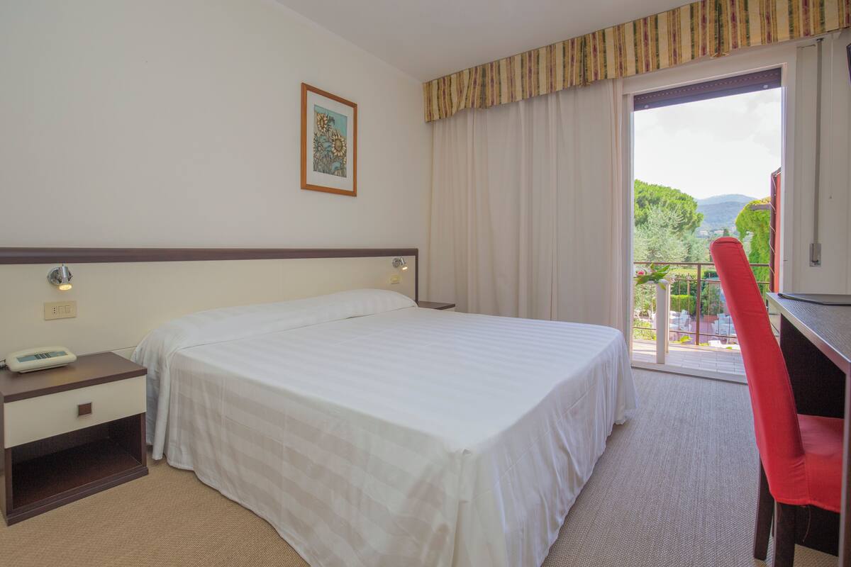 A hotel room with a bed, a desk, a chair, a window with curtains, and a painting on the wall.