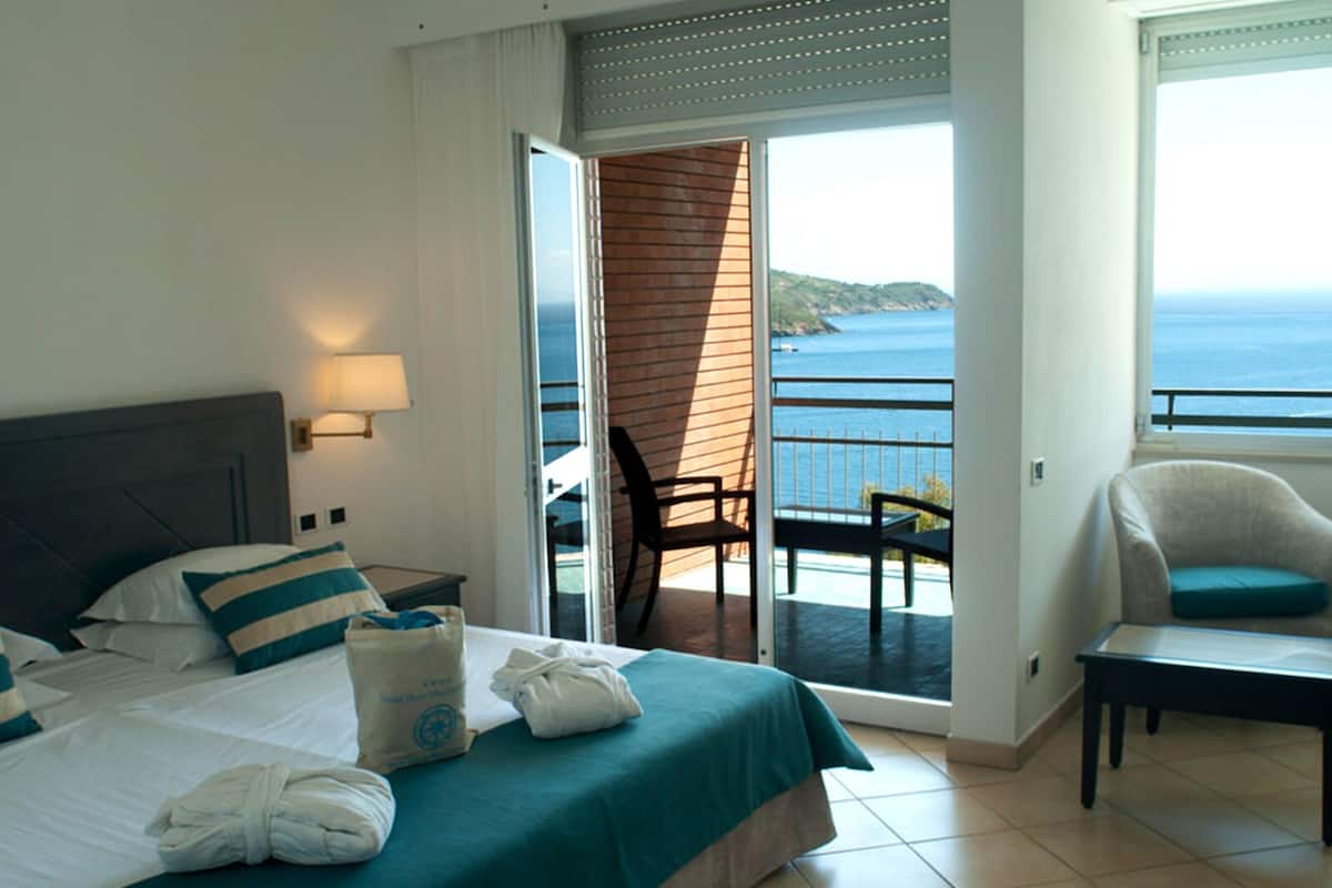 Habitación de hotel con cama, una silla, una mesa, un balcón con vista al mar y una ventana.