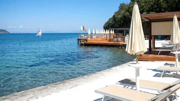 A beachside hotel with lounge chairs and umbrellas, a wooden deck, and a sailboat in the water. - JR Resort Elba International (Capoliveri)