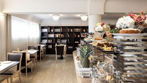 A modern dining area with wooden tables and chairs, a central buffet with various food items, and a bookshelf in the background. - Albergo Celide (Lucca)