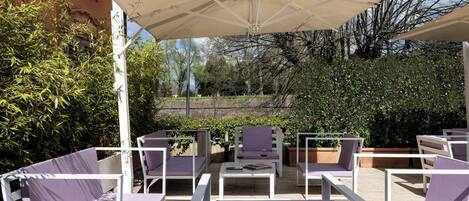 An outdoor seating area with white metal chairs and tables, covered by a large beige canopy.