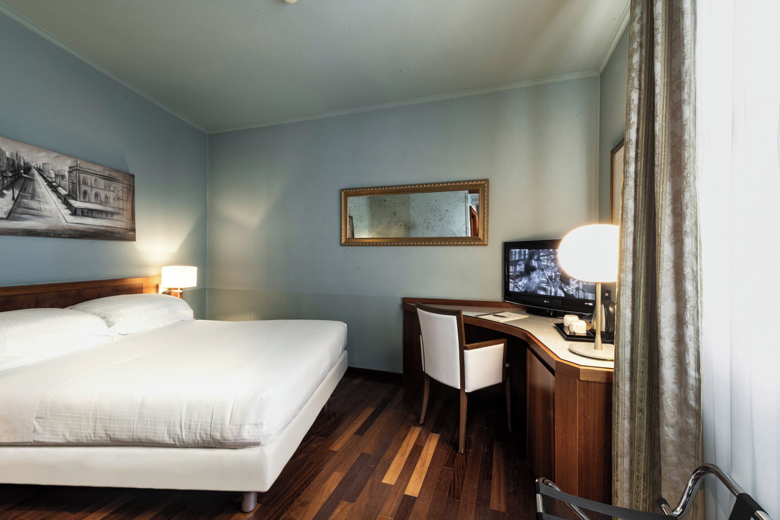 A hotel room with a bed, desk, chair, television, and a painting on the wall.