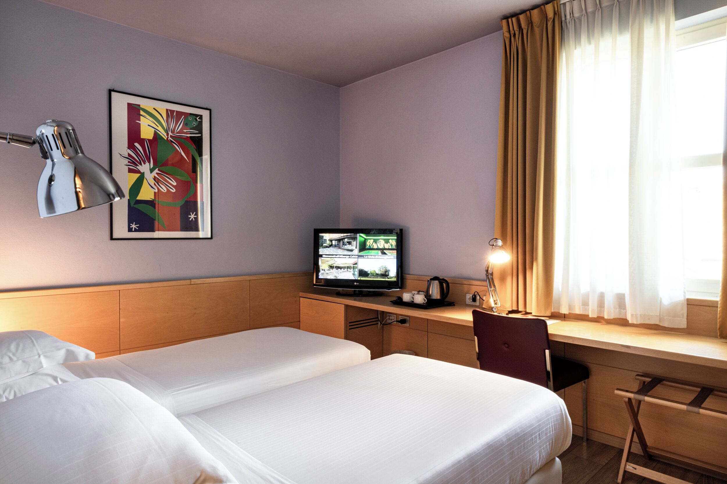 A hotel room with two beds, a desk with a computer, a television, and a window with curtains.
