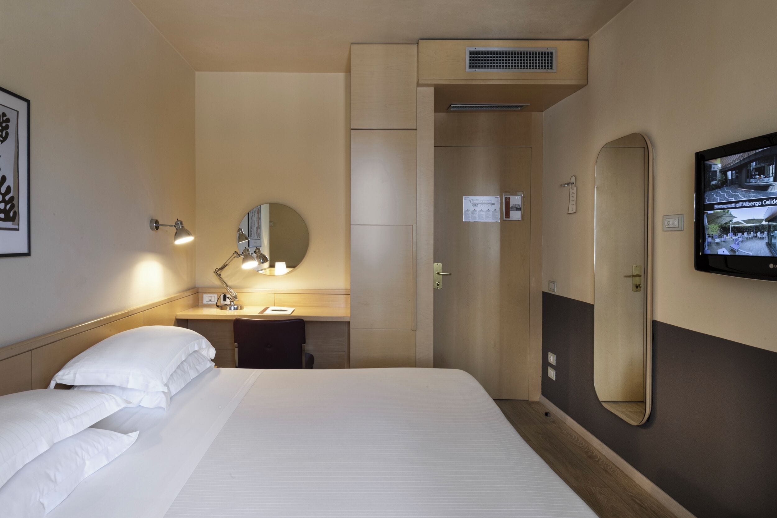 Habitación de hotel con una cama, un escritorio con lámpara, un espejo y un televisor en la pared.