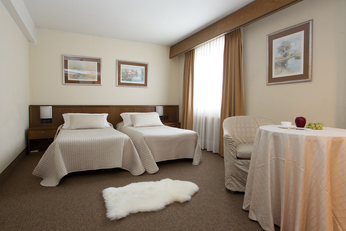 Ein Hotelzimmer mit zwei Betten, einem Esstisch mit einem roten Apfel und Trauben sowie zwei gerahmten Bildern an der Wand.