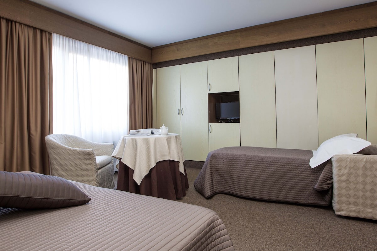 Ein Hotelzimmer mit zwei Betten, einem Sessel, einem Tisch mit Lampe und einem an der Wand befestigten Fernseher.
