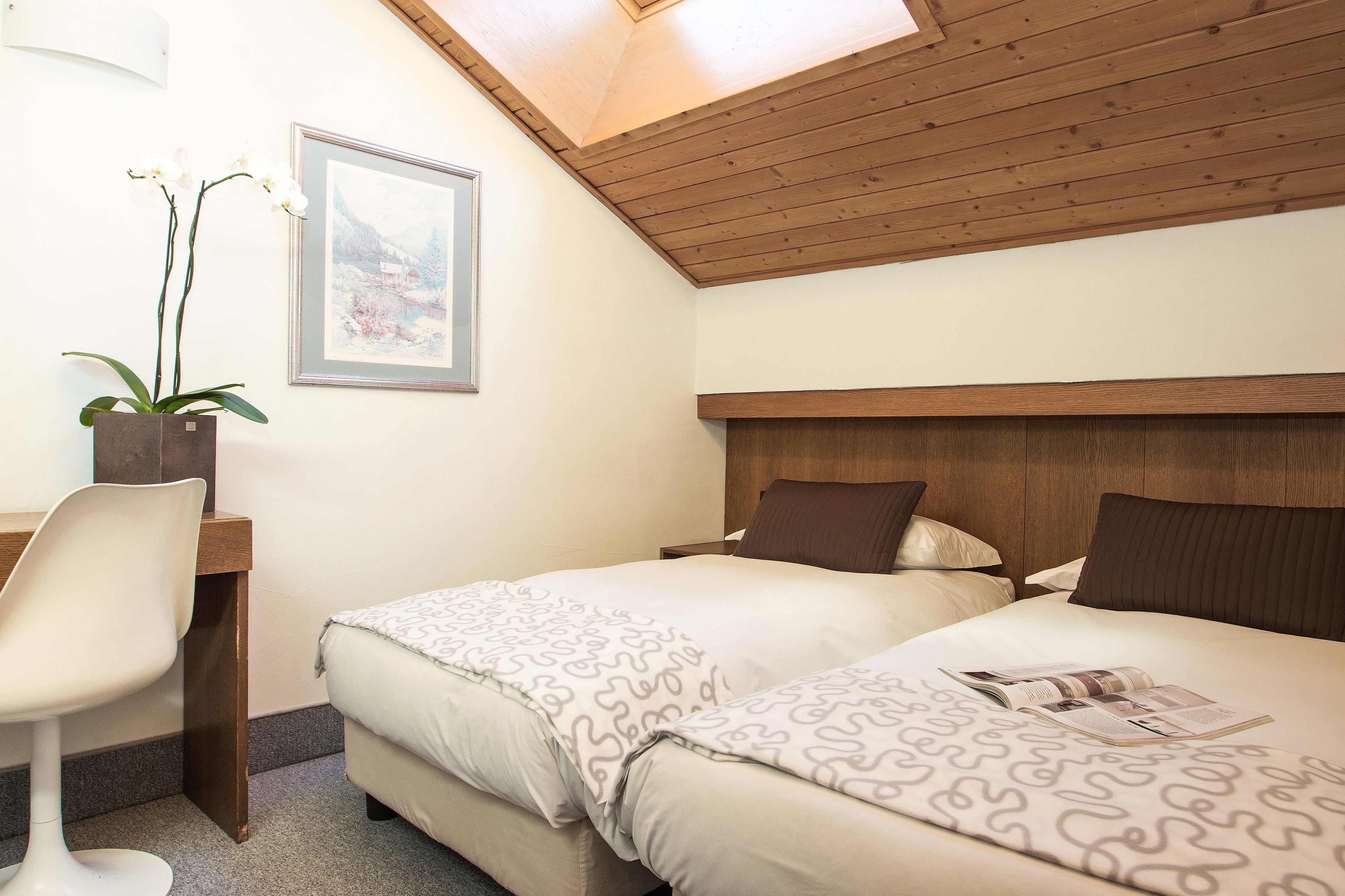 Ein Schlafzimmer mit Holzbalkendecke, zwei Betten mit gemusterten Betttextilien, einem Schreibtisch mit Stuhl und einem Topf mit einer Orchidee.