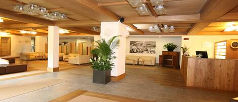 A spacious lobby with wooden flooring, a reception desk, seating areas, and a large potted plant.
