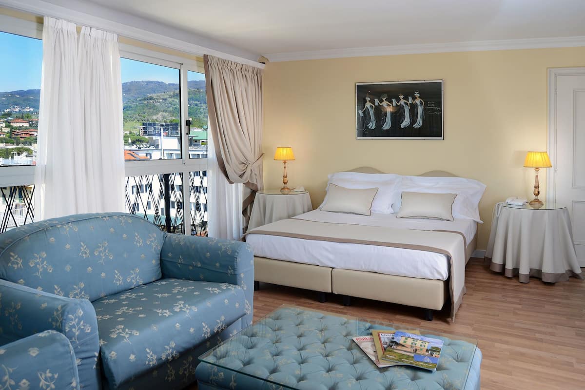 Quarto de hotel com cama, sofá, mesa de centro de vidro e vista para a cidade.
