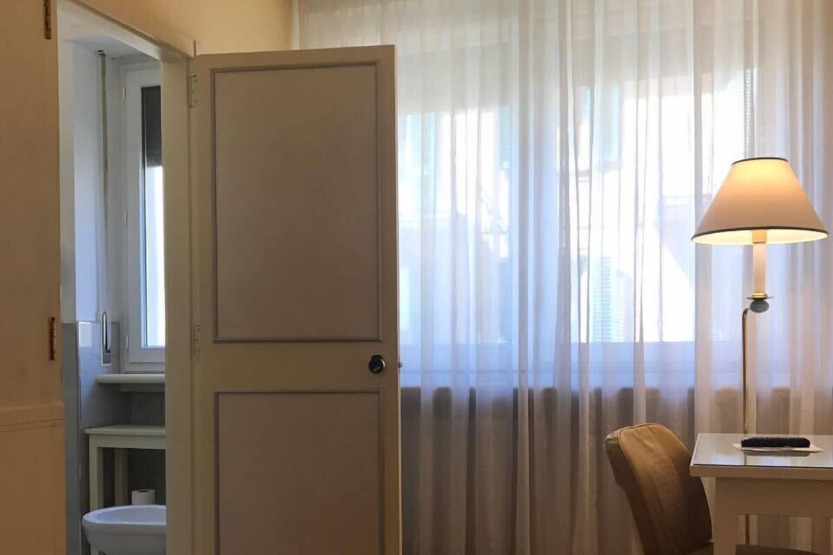 Quarto de hotel com cama, escrivaninha com luminária, cadeira, banheiro com vaso sanit ário e pia, e porta que dá para outro cômodo.