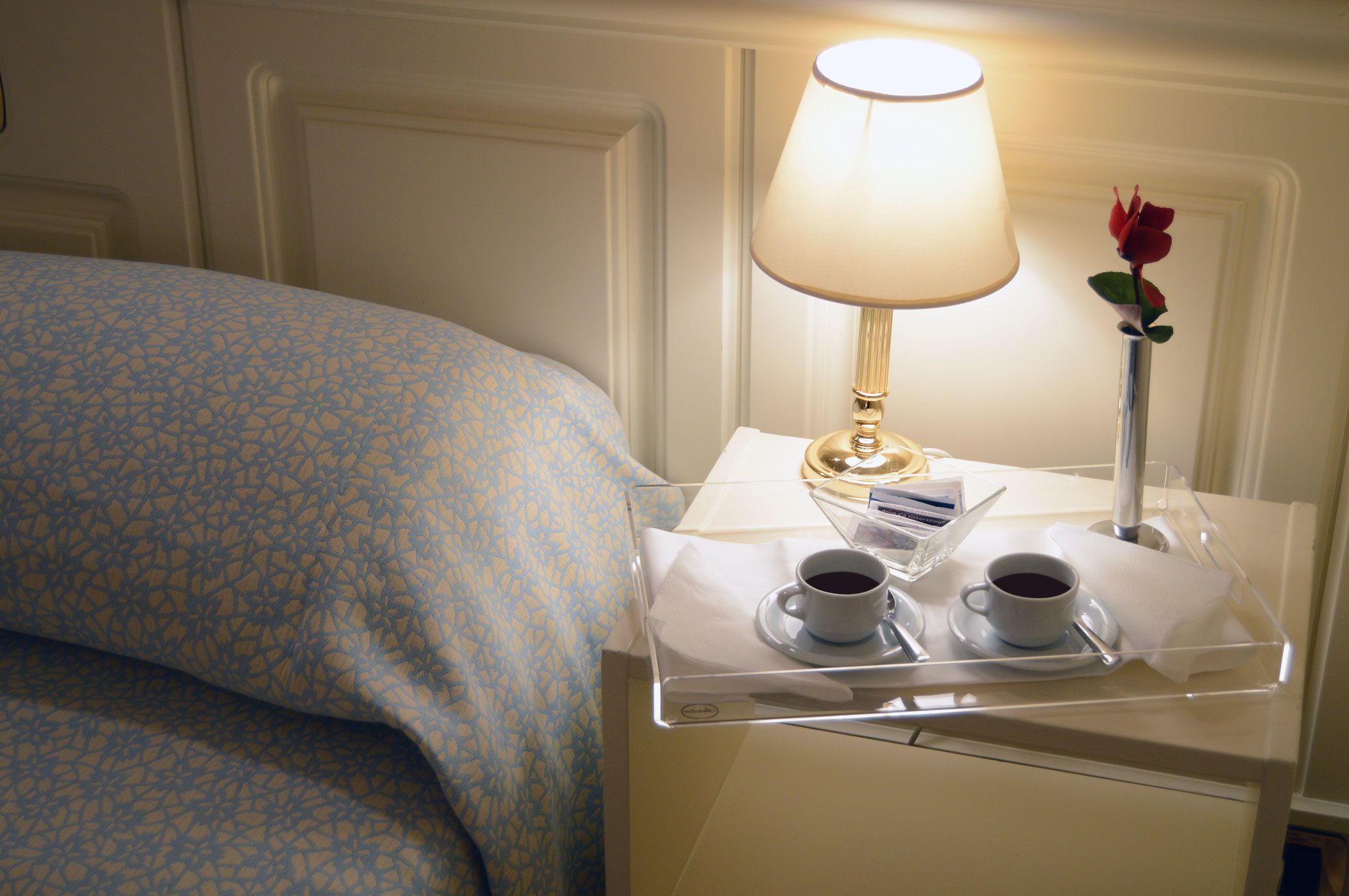 Ein ordentlich bezogenes Bett mit einer blauen, gemusterten Tagesdecke, ein Nachttisch mit Lampe, zwei Tassen Kaffee und eine Rose in einer Vase.