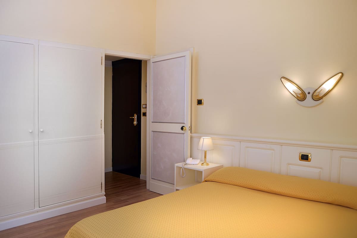 Quarto de hotel com cama, mesa de cabeceira com abajur, telefone e luminária na parede.