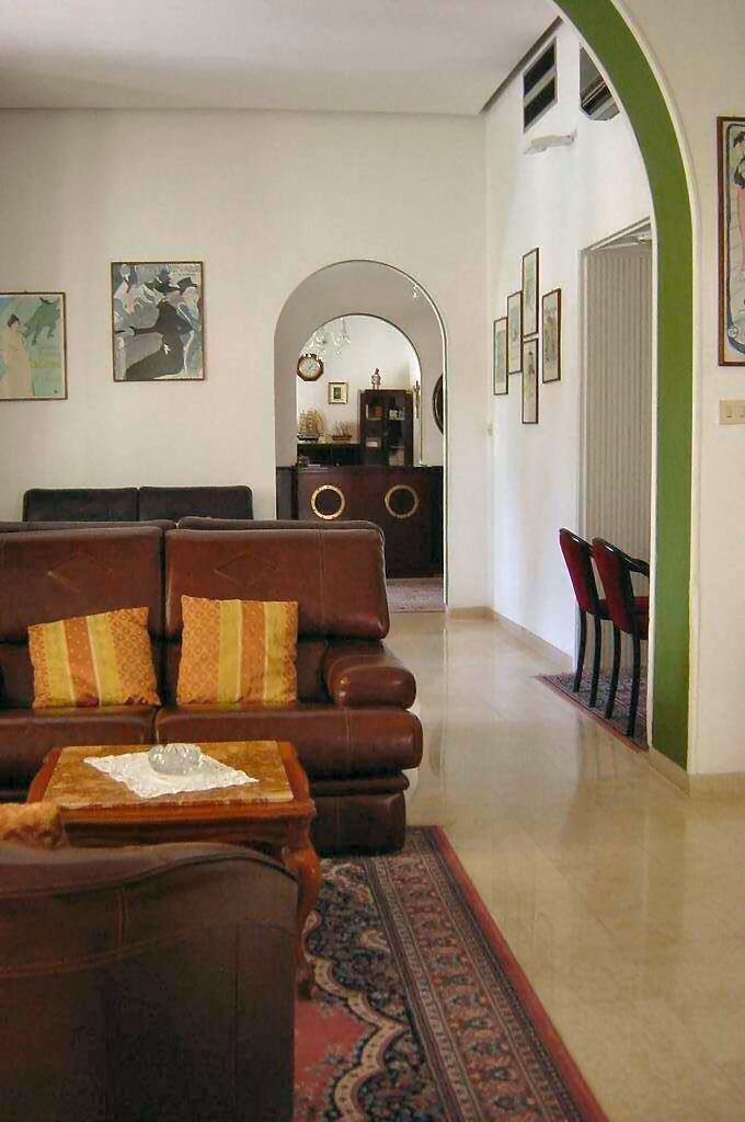 A living room with a brown sofa, a small table, and a patterned rug. There is a hallway with framed pictures on the walls and a door at the end.