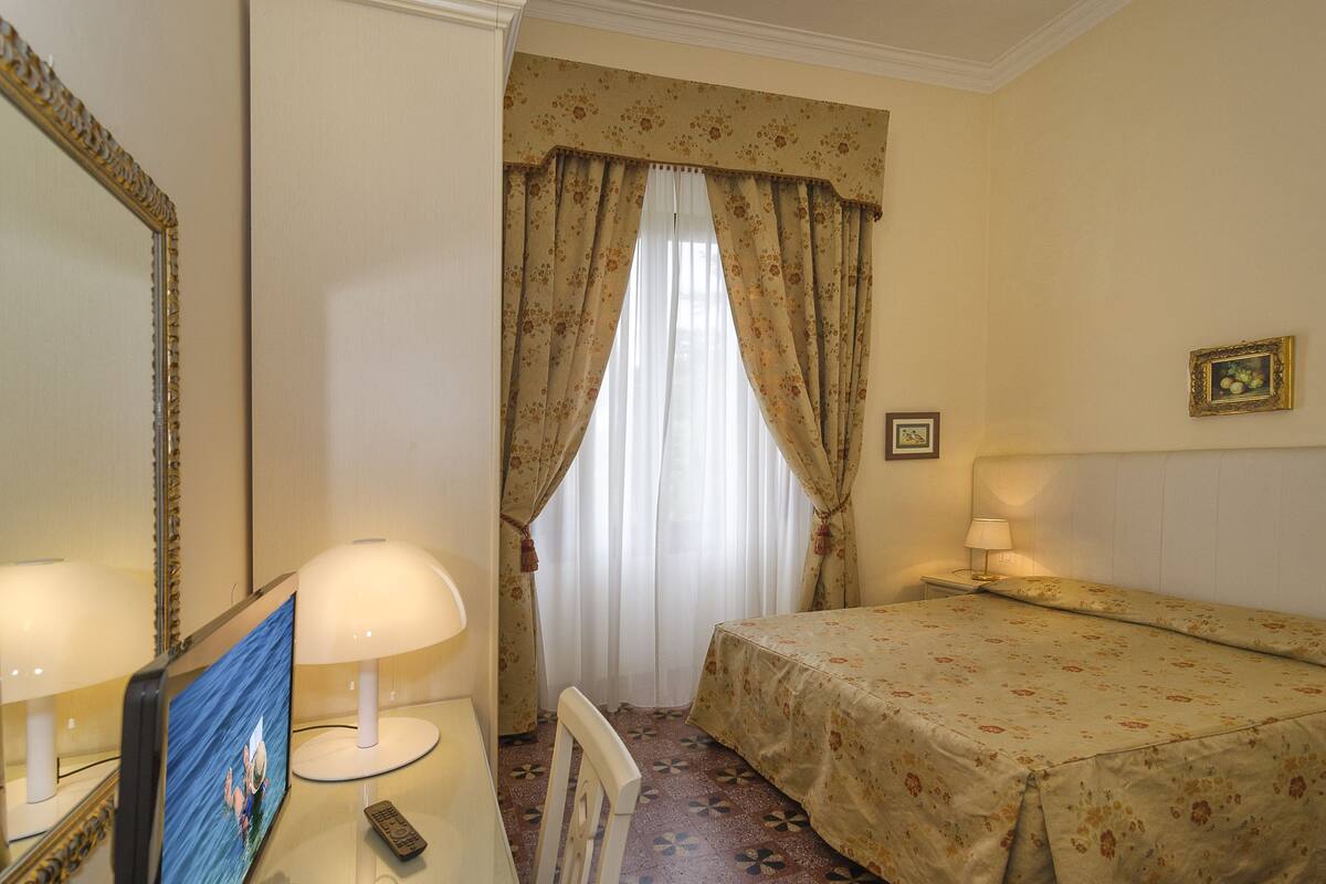 A hotel room with a bed, a desk with a lamp, a television, and a window with curtains.