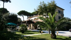 A hotel with a covered outdoor seating area, a well-maintained garden with palm trees, and a two-story building with balconies. - Hotel Verdemare (Pietrasanta)