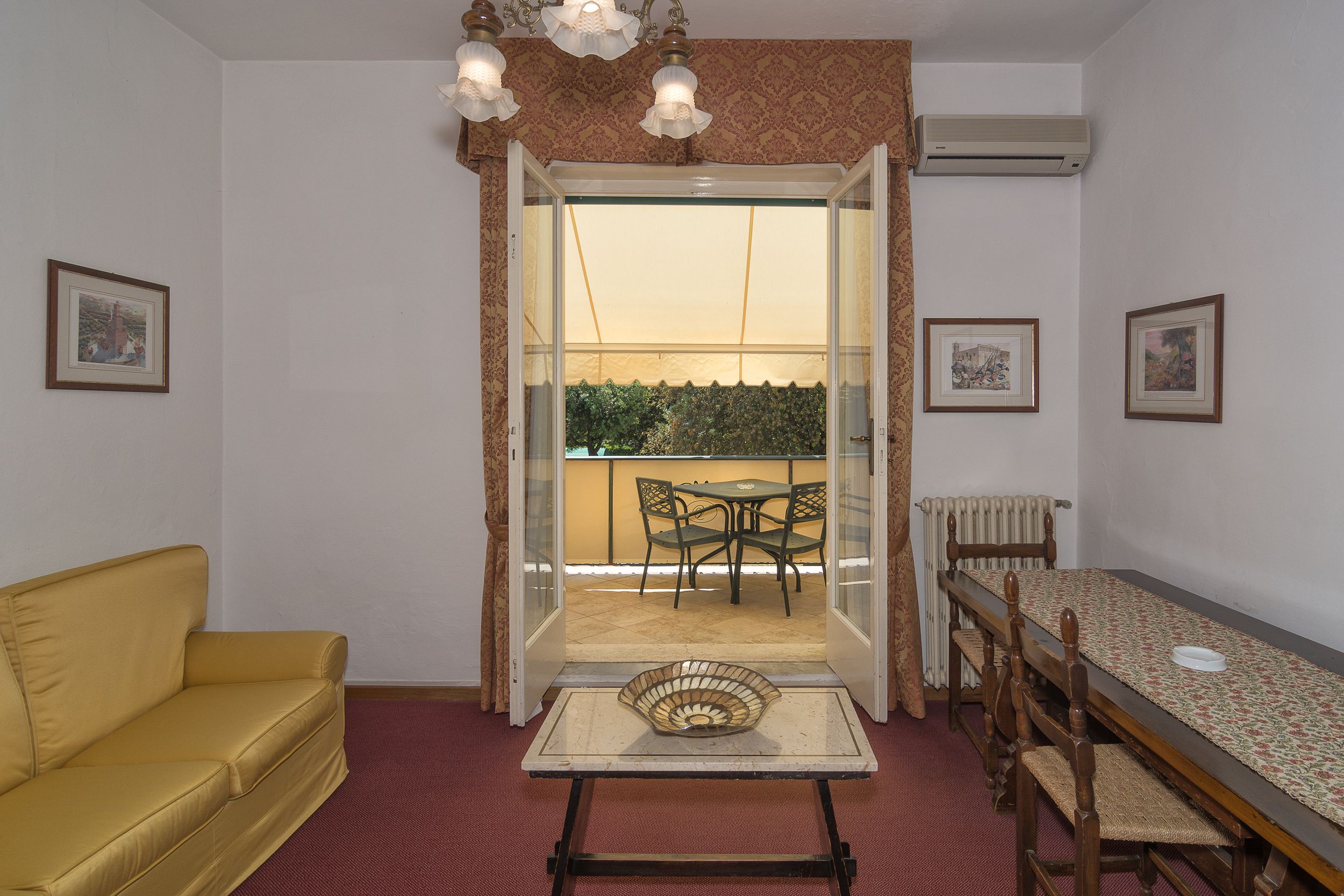 Une pièce avec un canapé jaune, une table à manger avec des chaises, et une vue sur un espace extérieur aménagé avec une table et des chaises, accessible par une porte vitrée.