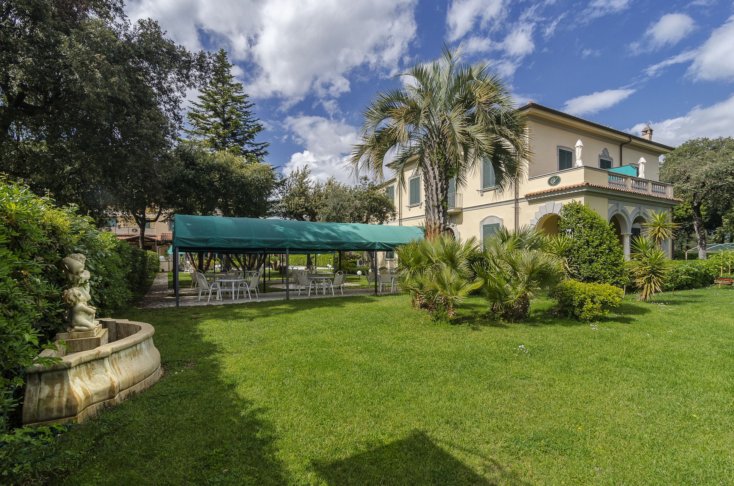 A well-maintained garden with a covered outdoor seating area, a fountain, and a large building with a balcony.
