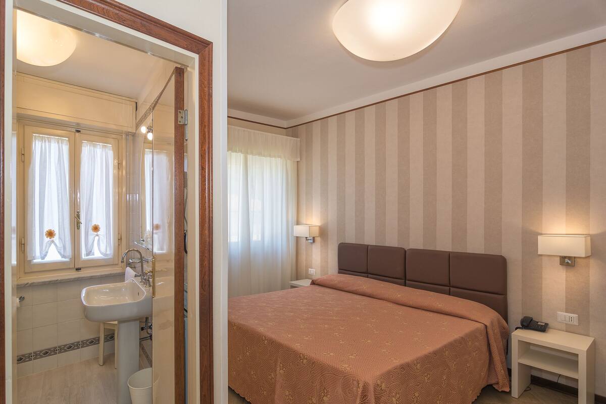 A hotel room with a bed, bedside table, lamp, and a bathroom with a sink and mirror.