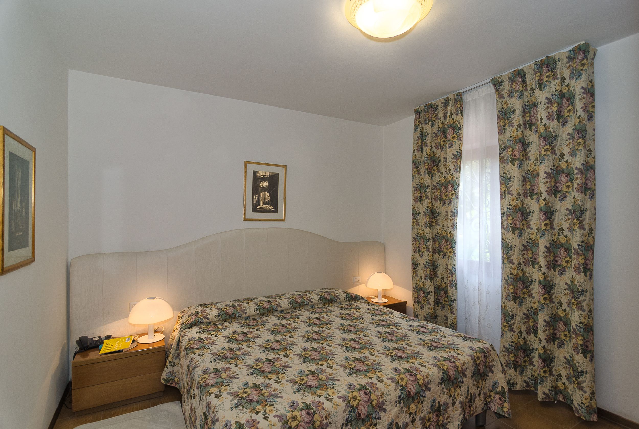 A bedroom with a bed, two bedside tables with lamps, a framed picture on the wall, and floral curtains.