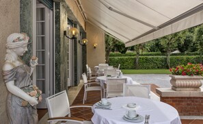 An outdoor patio with white tables and chairs, a statue of a woman holding a flower, and a garden view. - Hotel Villa Edera (Camaiore)