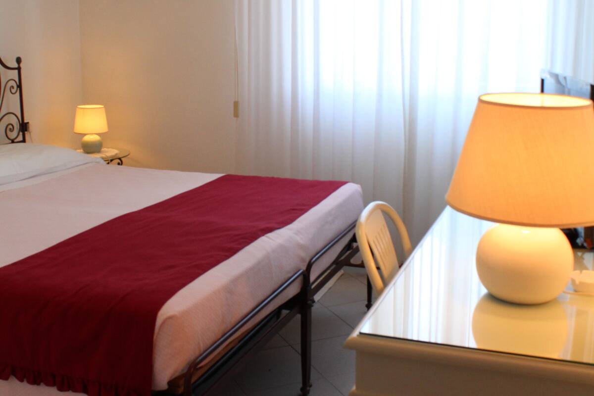 A hotel room with a bed, a table with a lamp, a chair, a nightstand with a lamp, and a window with curtains.