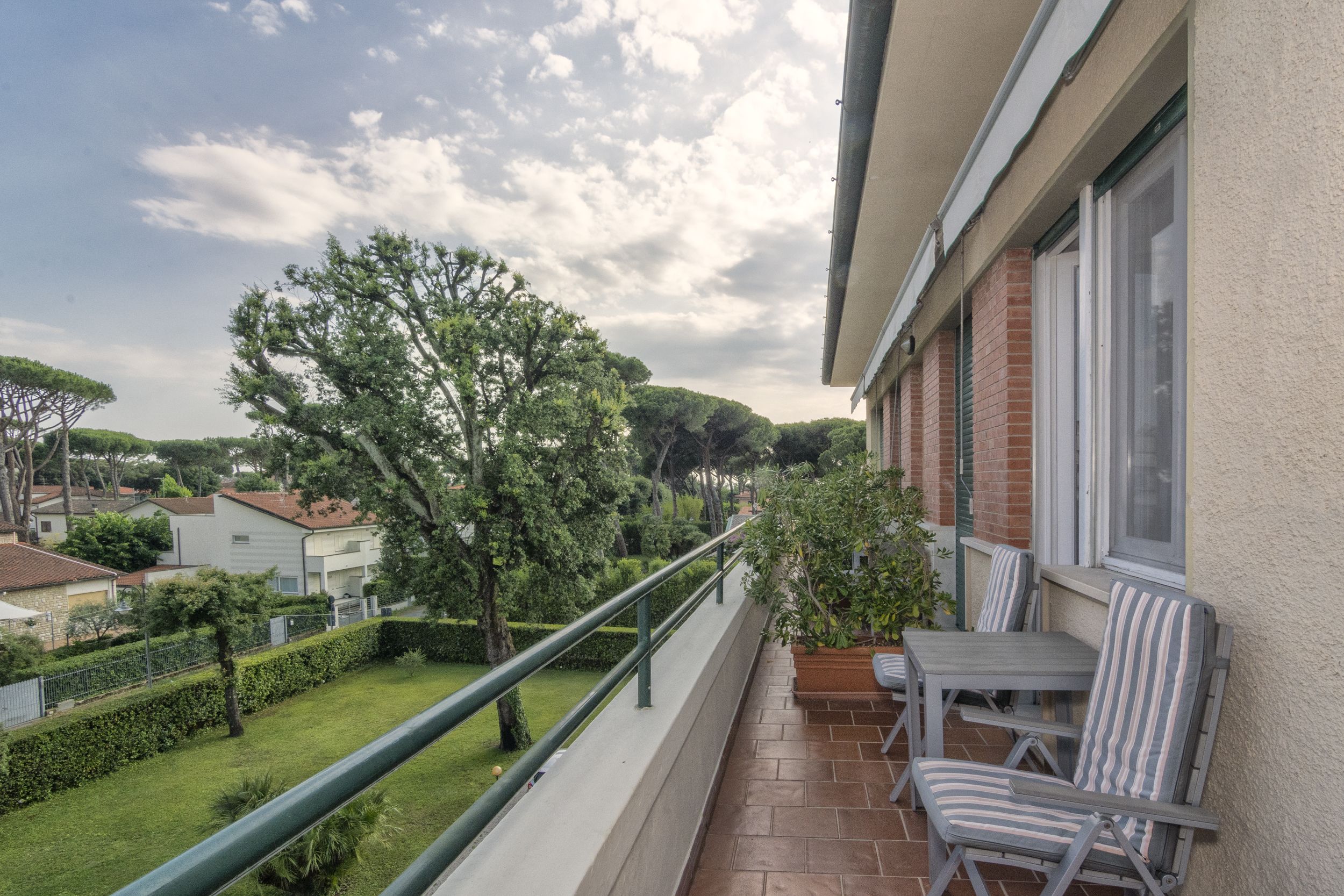 A balcony with a view of a well-maintained garden, a table and chairs, and a railing.