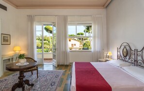 A hotel room with a bed, a table with a plant, a chair, and a view of greenery outside. - Hotel Villa Edera (Camaiore)