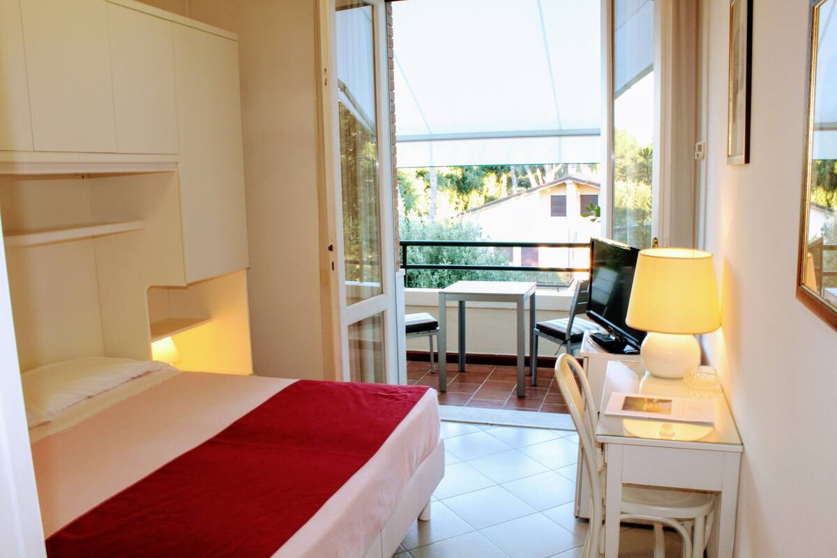 A hotel room with a bed, a desk with a lamp, and a view of the outside balcony.