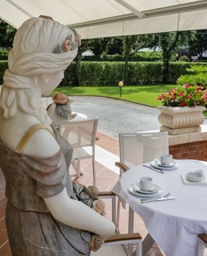 A statue of a woman in a dress seated at a table with a white tablecloth, surrounded by chairs, in an outdoor setting with a garden and a hedge in the background. - Hotel Villa Edera (Camaiore)