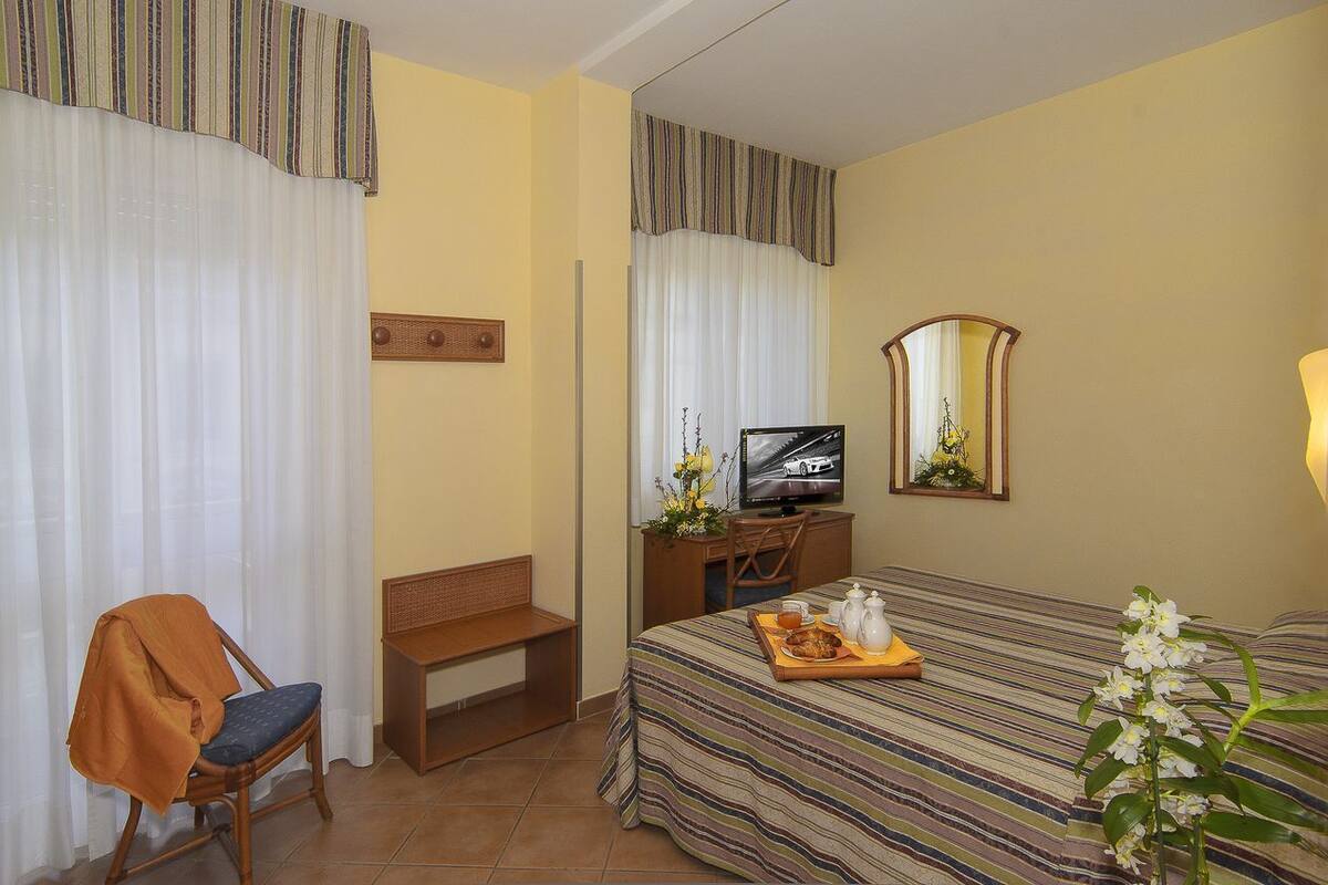 A hotel room with a bed, a desk with a television, a chair, a bench, and a tray with breakfast items.