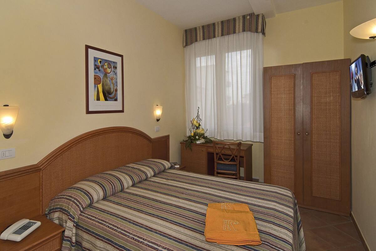 A hotel room with a bed, bedside table, lamp, artwork, window, and wardrobe.