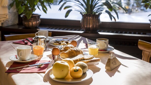 A breakfast table with a variety of food items, a window with a view of trees, and a potted plant on the windowsill.