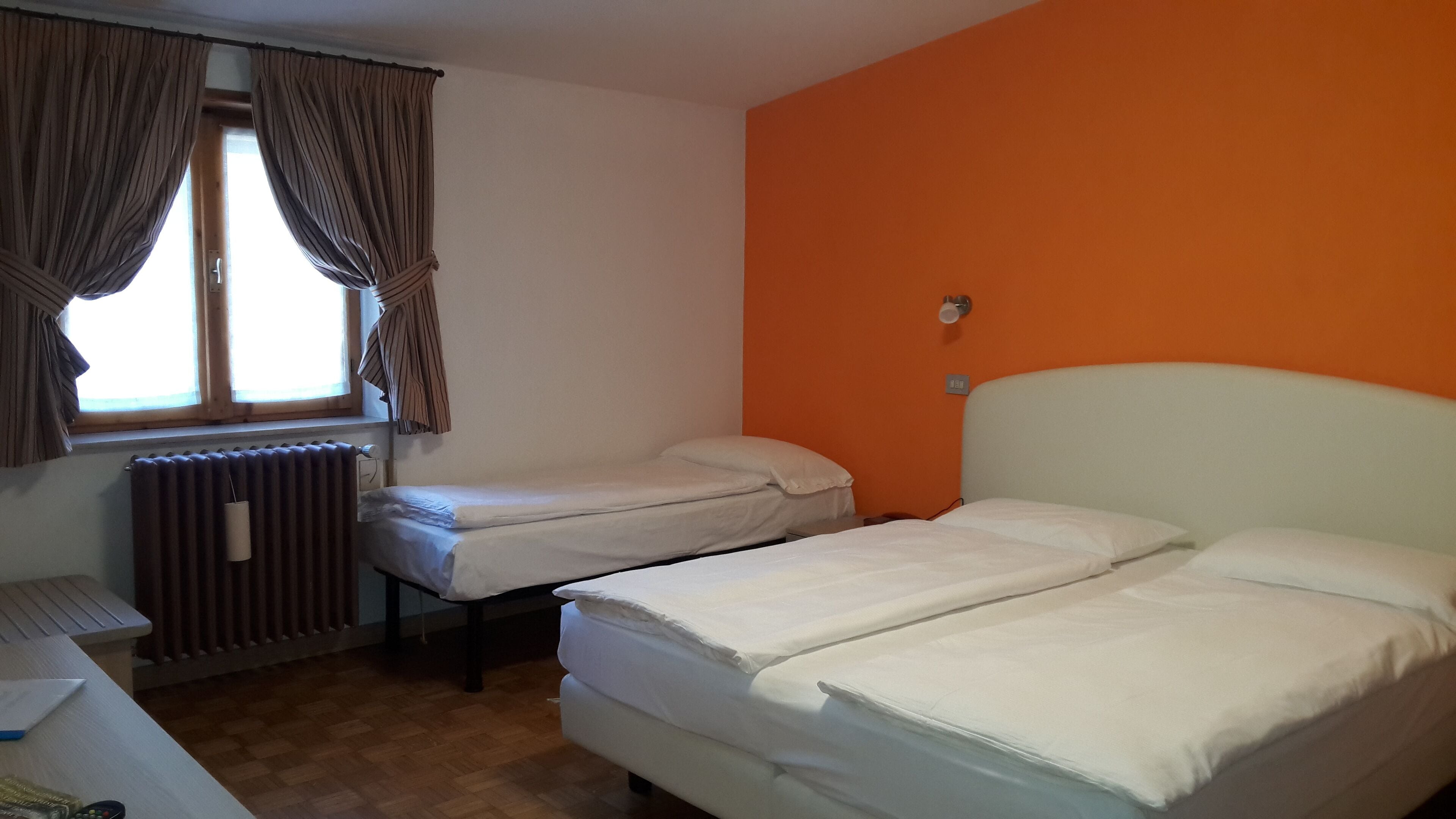 A room with two beds, a radiator, a window with curtains, and an orange wall.
