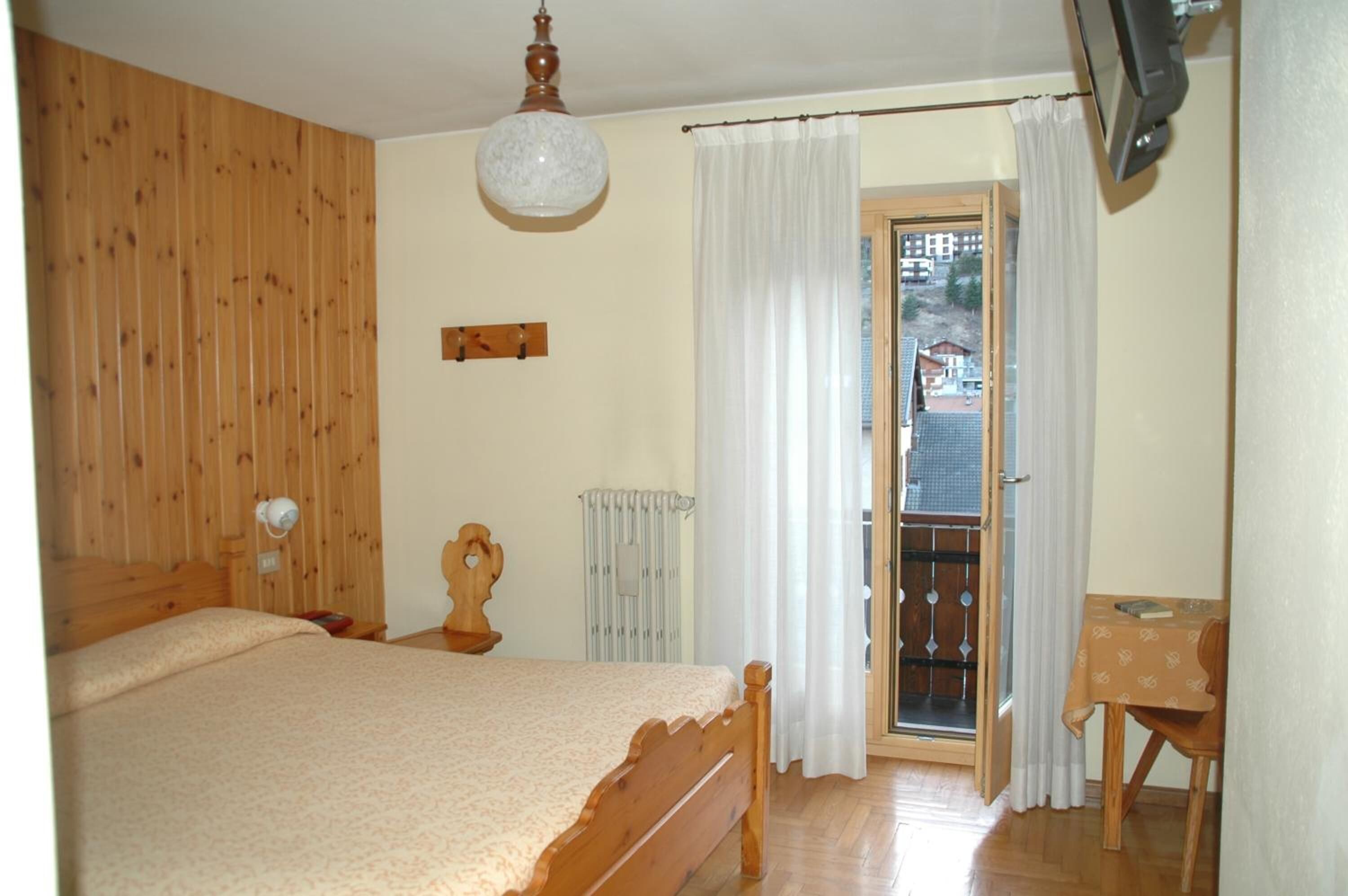 Un dormitorio con una cama de madera, una lámpara en el techo, un radiador, una mesita y vistas a través de una puerta.
