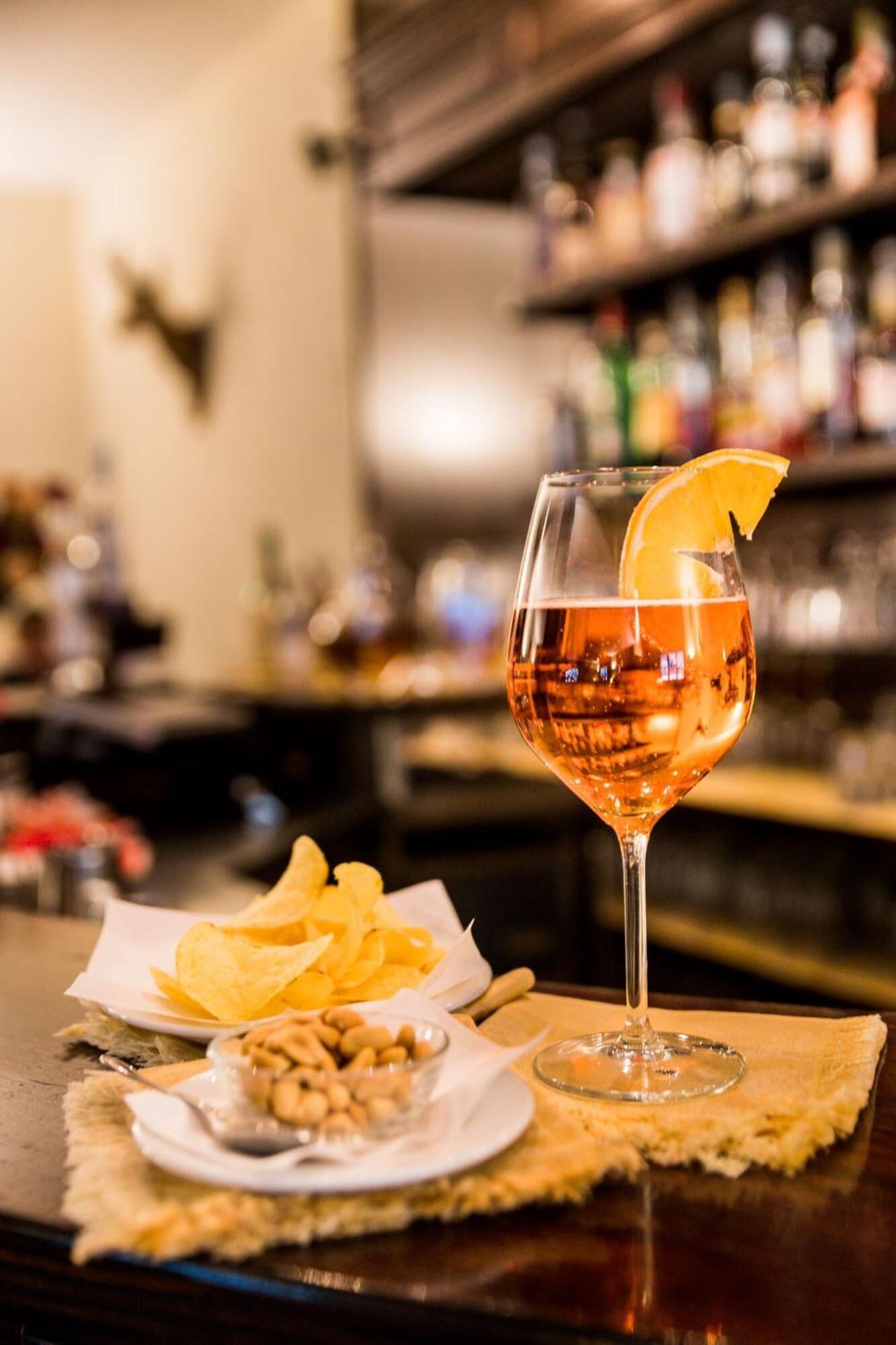 A cocktail glass with an orange slice garnish on a bar counter, accompanied by a plate of chips and a small bowl of nuts.