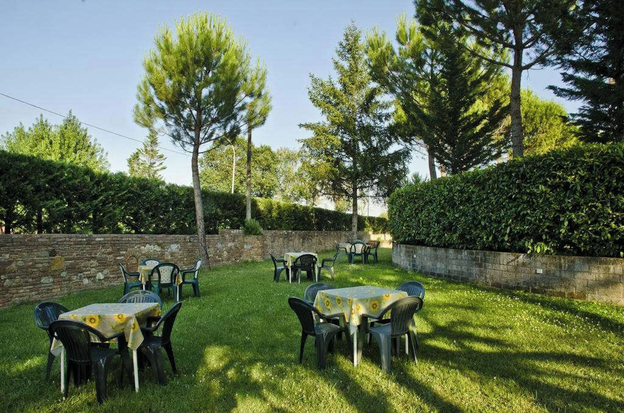 An outdoor seating area with tables and chairs, surrounded by trees and a stone wall.