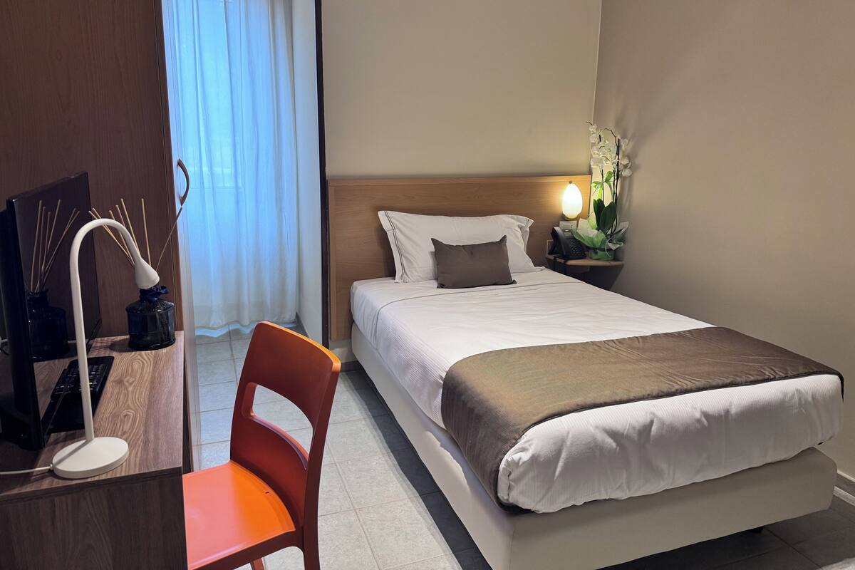A hotel room with a bed, a desk with a chair, a lamp, a telephone, and a plant.