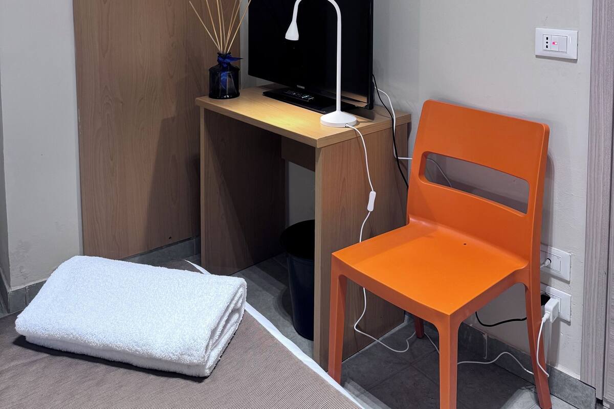 A small, neatly organized room with a wooden door, a desk with a computer and a lamp, an orange chair, and a white towel on the floor.