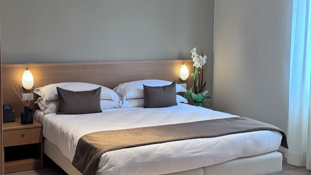 A neatly made bed with white and brown bedding, a wooden headboard, bedside lamps, and a vase with white flowers. - Hotel La Magione (Poggibonsi)