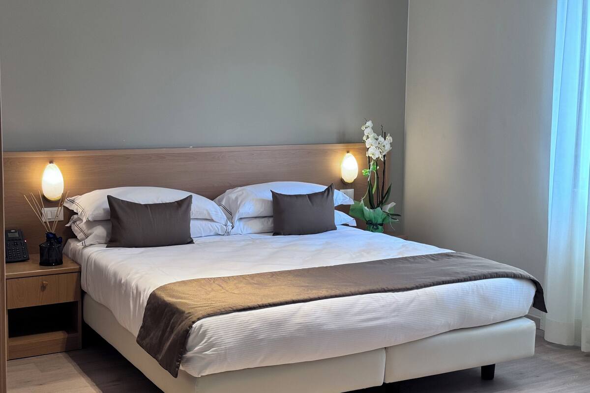 A neatly made bed with white and brown bedding, a wooden headboard, bedside lamps, and a vase with white flowers.
