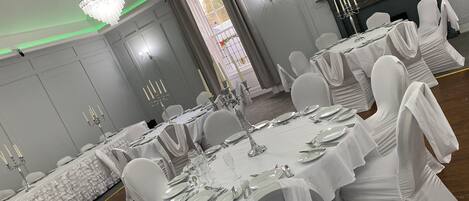 Une salle de banquet avec des tables rondes dressées pour le repas, des chaises blanches et des lustres.