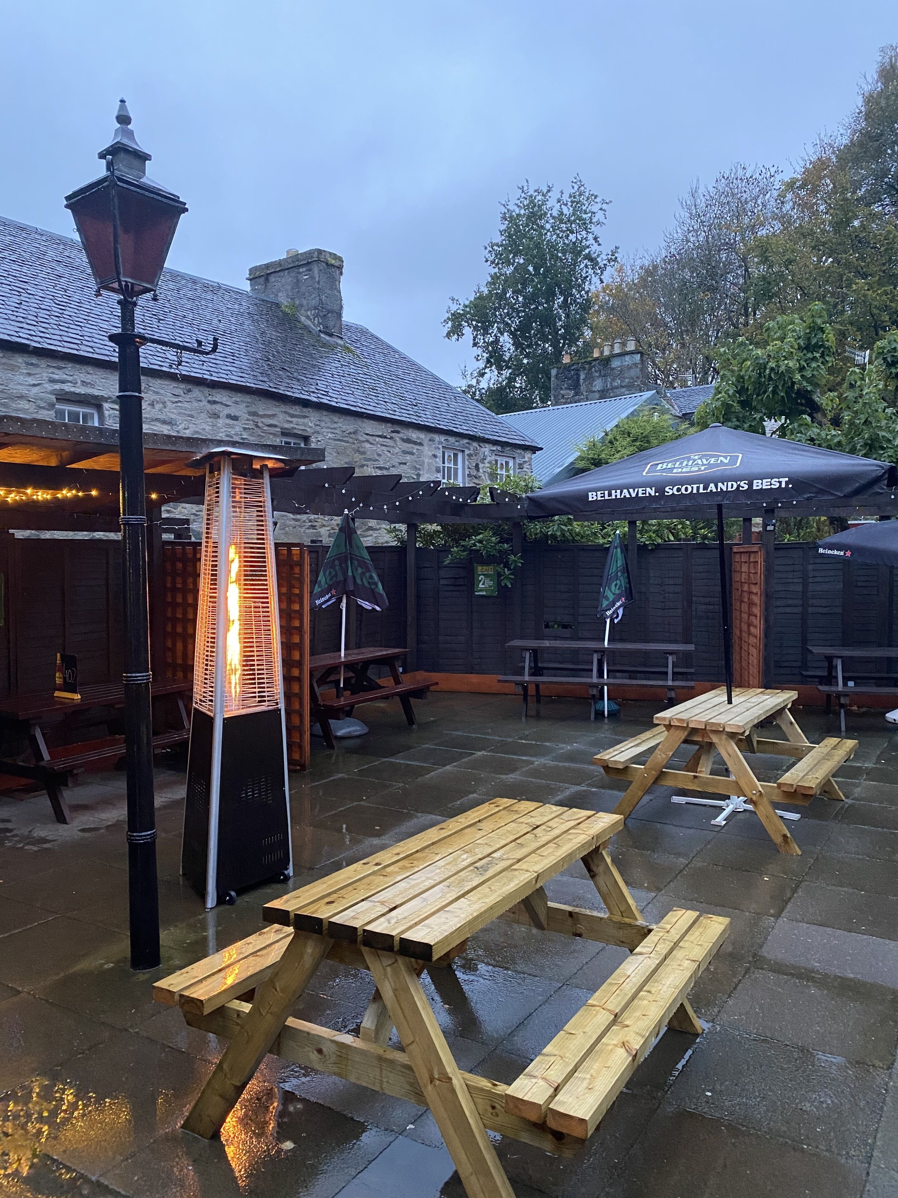 An outdoor seating area with wooden tables and benches, a stone building in the background, and a sign for Belhaven Brewery.