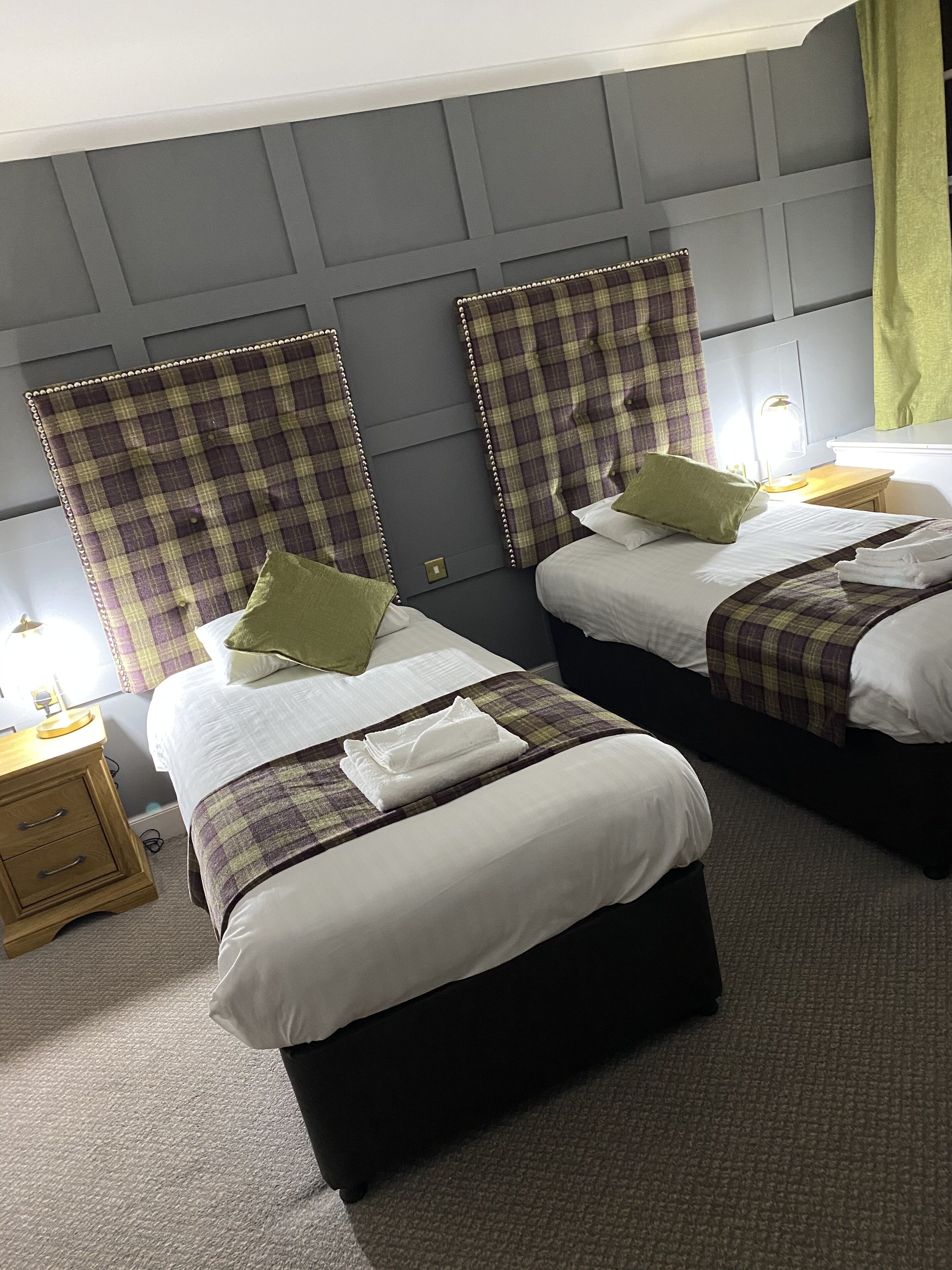 A hotel room with two beds, each with a plaid headboard, white bedding, and a green pillow. There is a wooden nightstand with a lamp and a power outlet.