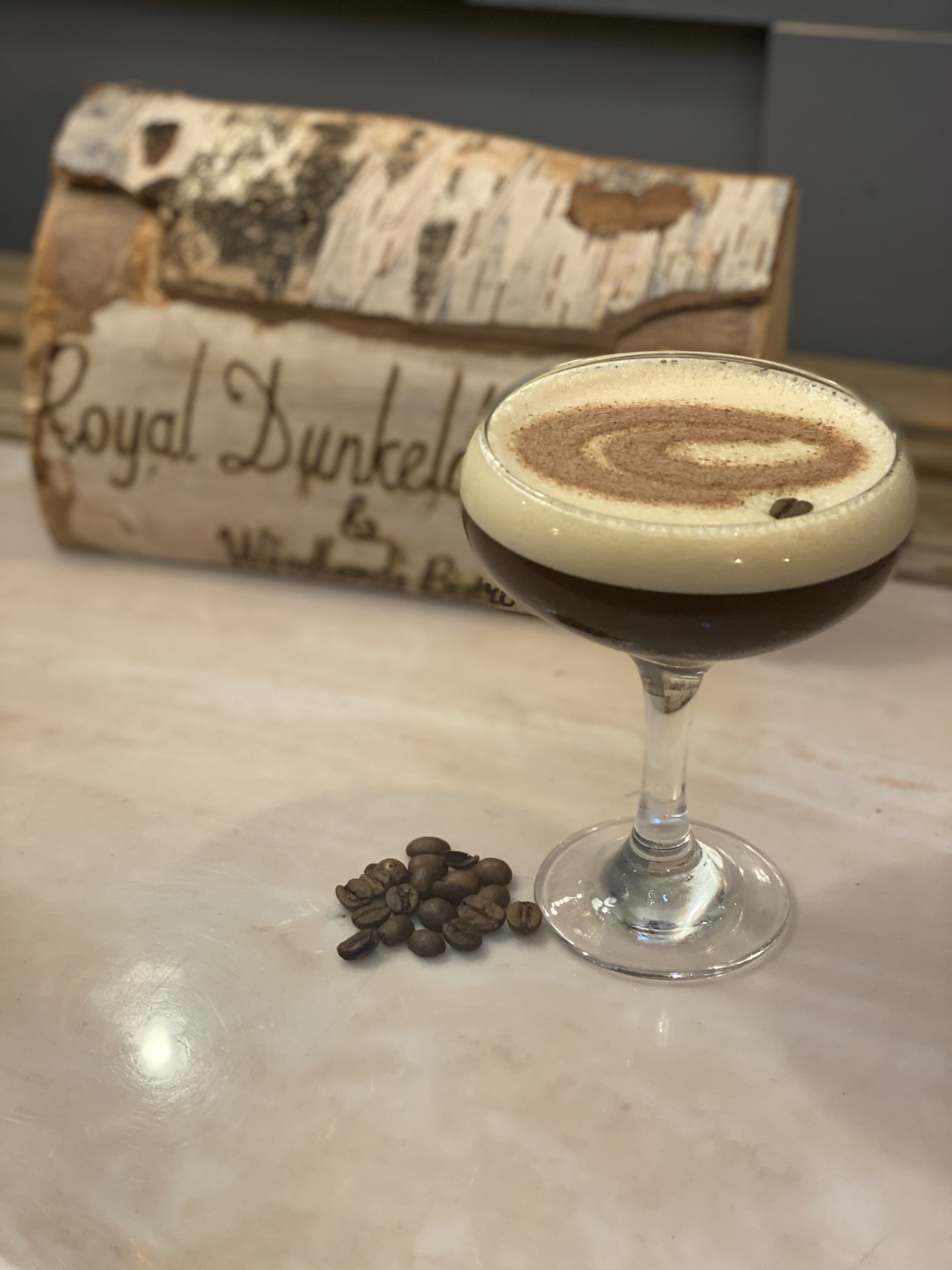 Een cocktail in een martini-glas met een spiraalmotief, koffiebonen en een rustiek houten bordje op de achtergrond.