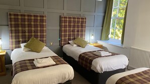 A hotel room with three beds, each with a plaid bedspread and pillows, a window with a view of greenery, and a radiator on the wall. - Royal Dunkeld (Dunkeld)
