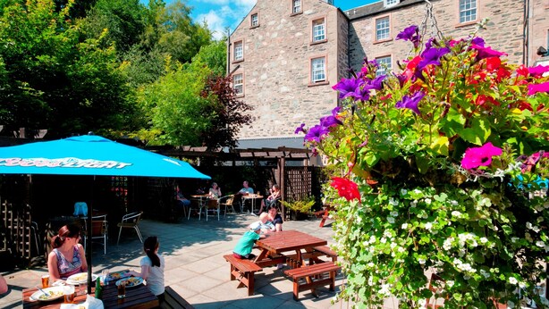 An outdoor patio area with stone buildings, wooden tables, and benches. - Royal Dunkeld (Dunkeld)
