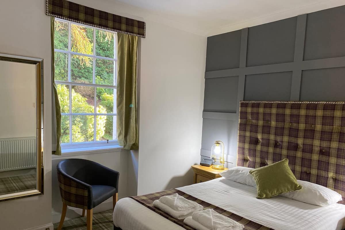 A hotel room with a bed, a chair, a bedside table with a lamp, and a window with curtains.